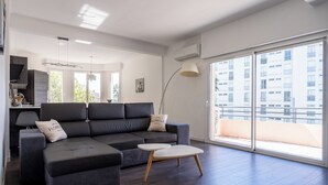 Uma sala de estar moderna com um sofá seccional cinza, uma pequena mesa de centro branca e uma grande porta de vidro deslizante que dá para uma varanda.