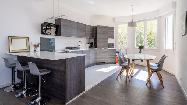 Uma cozinha moderna com uma ilha central, banquetas, uma mesa de jantar com cadeiras e grandes janelas que oferecem luz natural.