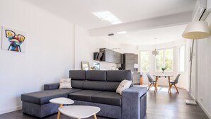 Uma sala de estar moderna com um sofá seccional cinza, duas mesas laterais brancas e uma área de jantar com uma mesa redonda e cadeiras.