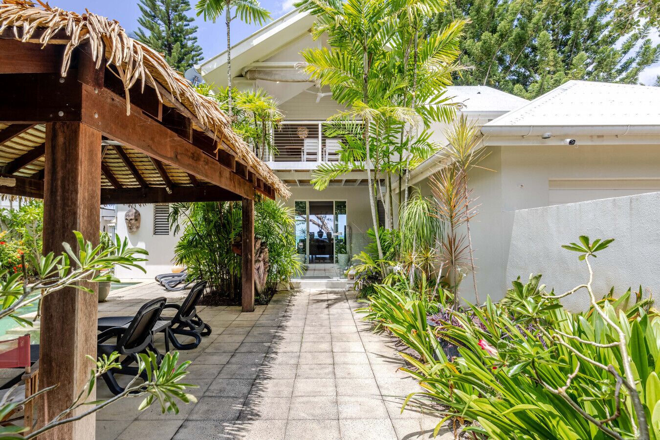 A covered outdoor seating area with chairs, surrounded by lush tropical plants and a pathway leading to a building entrance.