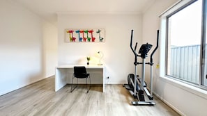 A room with a white desk, a black chair, a standing desk lamp, a treadmill, and a colorful giraffe painting on the wall. - Tarneit Holiday Home (Tarneit)