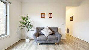 A living room with a grey armchair, a white cushion, and a potted plant. There are two framed pictures on the wall and a vent on the right wall. - Tarneit Holiday Home (Tarneit)