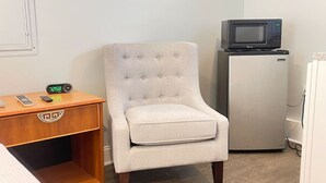 A small room with a light-colored armchair, a wooden nightstand with a clock, a microwave on a small fridge, and a bed with a white headboard.