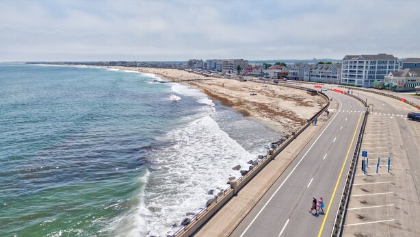 A coastal road with a beach on one side and buildings in the distance.