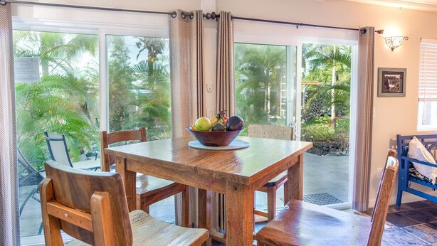 A wooden dining table with a bowl of fruit, surrounded by chairs, in a room with large glass doors leading to a tropical garden. - Sunrise Tropical Garden Getaway (Pahoa)