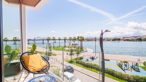 A balcony with a view of a waterfront promenade, including a bridge, palm trees, and a swimming area. - Welcomely - Superior Room - Dith Piaf (Olbia)