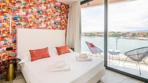 Habitación de hotel con una cama grande, dos almohadas rojas, un cabecero blanco y vistas a un cuerpo de agua y un paisaje urbano a través de amplios ventanales.