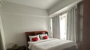 A bedroom with a wooden bed, white bedding, and red pillows. There is a bedside table with a lamp and a small plant. The room has white walls and a window with curtains. - 150 Newport condotel pasay  (Pasay)
