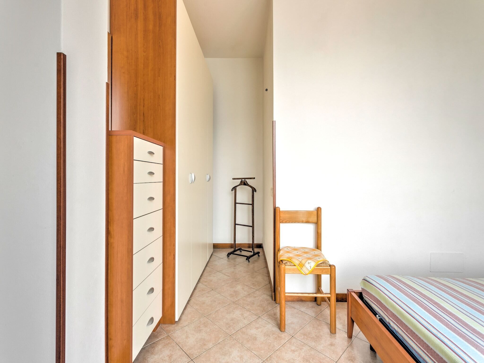 A narrow room with a wooden wardrobe, a chair, a bed, and a trolley.