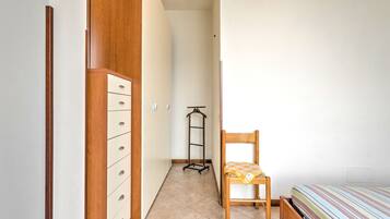 A narrow room with a wooden wardrobe, a chair, a bed, and a trolley.