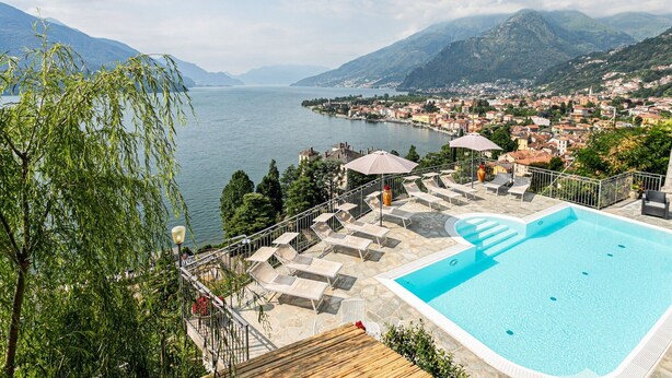 Piscina con sillones de descanso y sombrillas, con vistas a un lago y una ciudad con montañas al fondo.