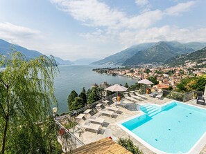 A swimming pool with lounge chairs and umbrellas, overlooking a lake and a town with mountains in the background.
