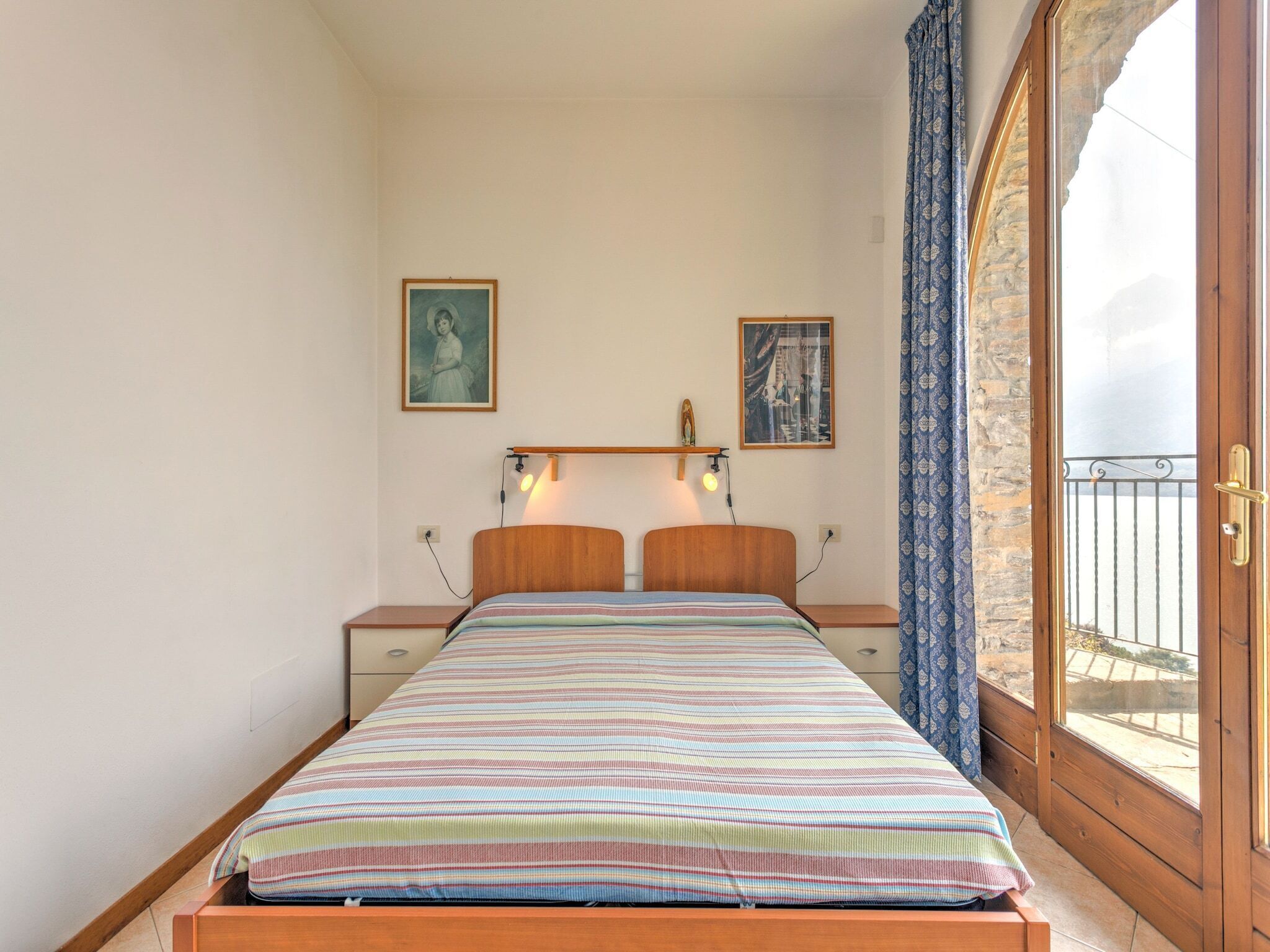 Un dormitorio con una cama de madera, dos mesitas de noche, una lámpara auxiliar y una ventana con vistas, además de dos cuadros en la pared.