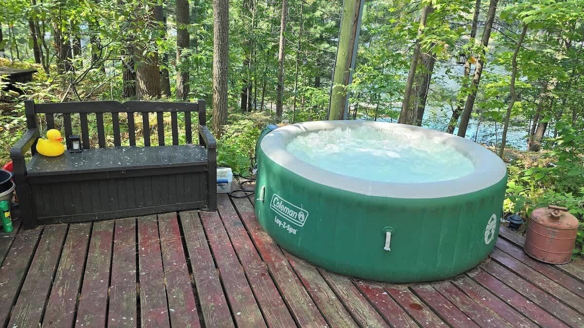 A hot tub on a wooden deck surrounded by trees, a bench, and a rubber duck.
