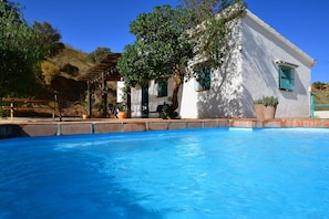 Vue sur une piscine, avec en arrière-plan un bâtiment blanc aux volets verts, entouré de plantes en pot et surmonté d’une pergola.