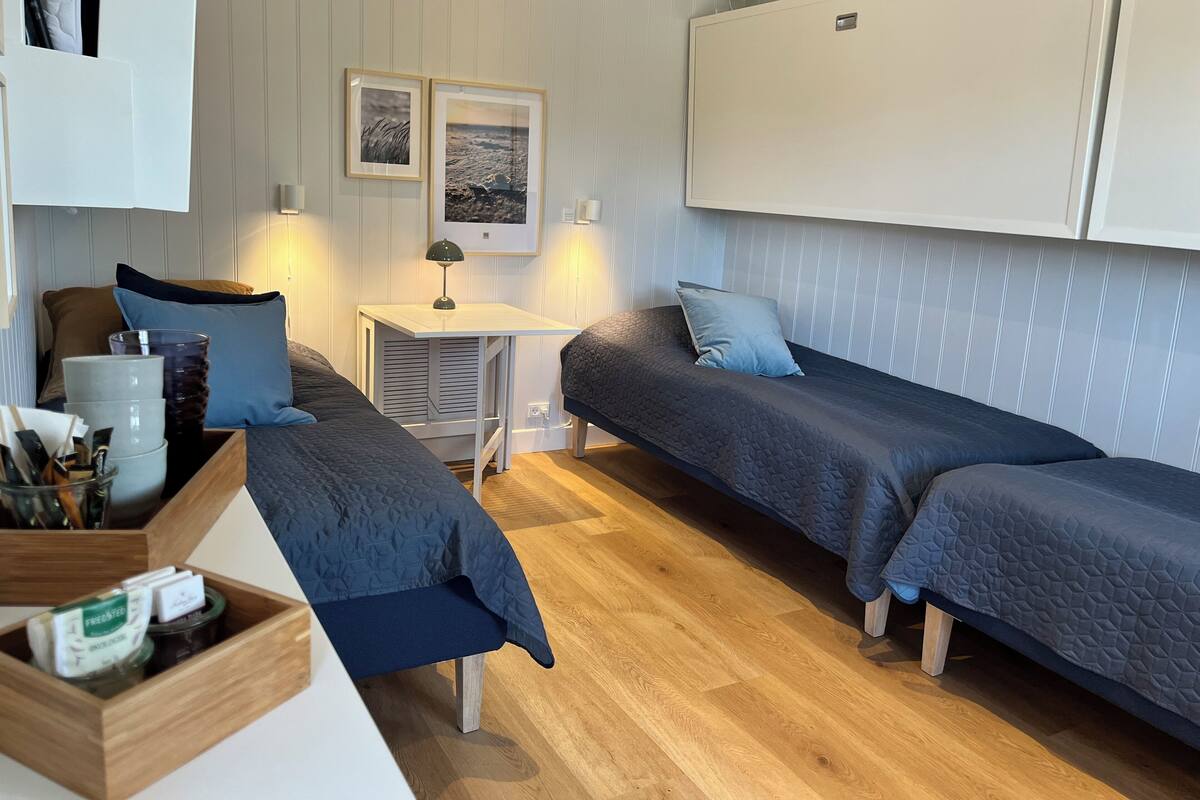 A compact room with two single beds, a small table, and a bench. There are framed pictures on the wall and a white cabinet above the bed.