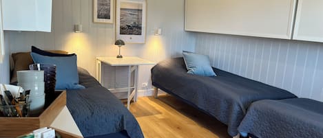 A compact room with two single beds, a small table, and a bench. There are framed pictures on the wall and a white cabinet above the bed.