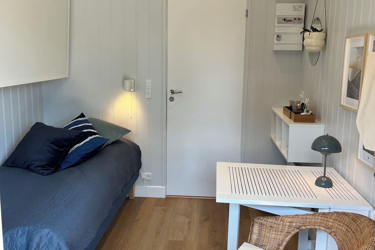 A compact room with a bed, a small table with a lamp, a wicker chair, a white door, and a wall-mounted air conditioning unit.