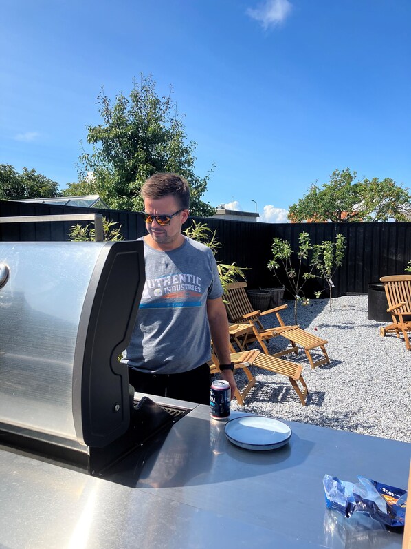 A man standing next to a barbecue grill with a can of beer on a table. - GuestHouse Læsø (Læsø)