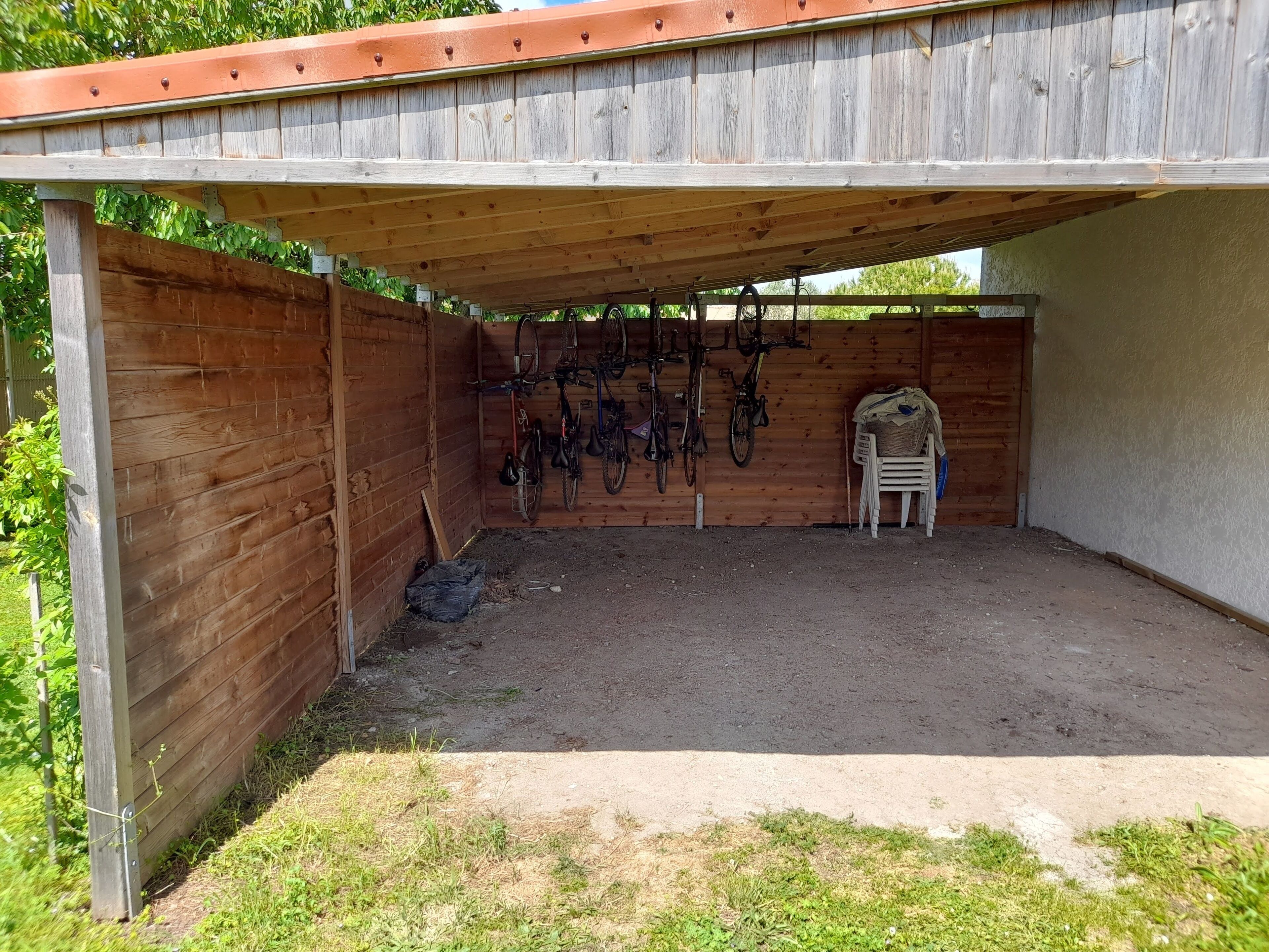 Un'area esterna coperta con struttura in legno, pavimento in terra battuta e diverse biciclette appese al muro.