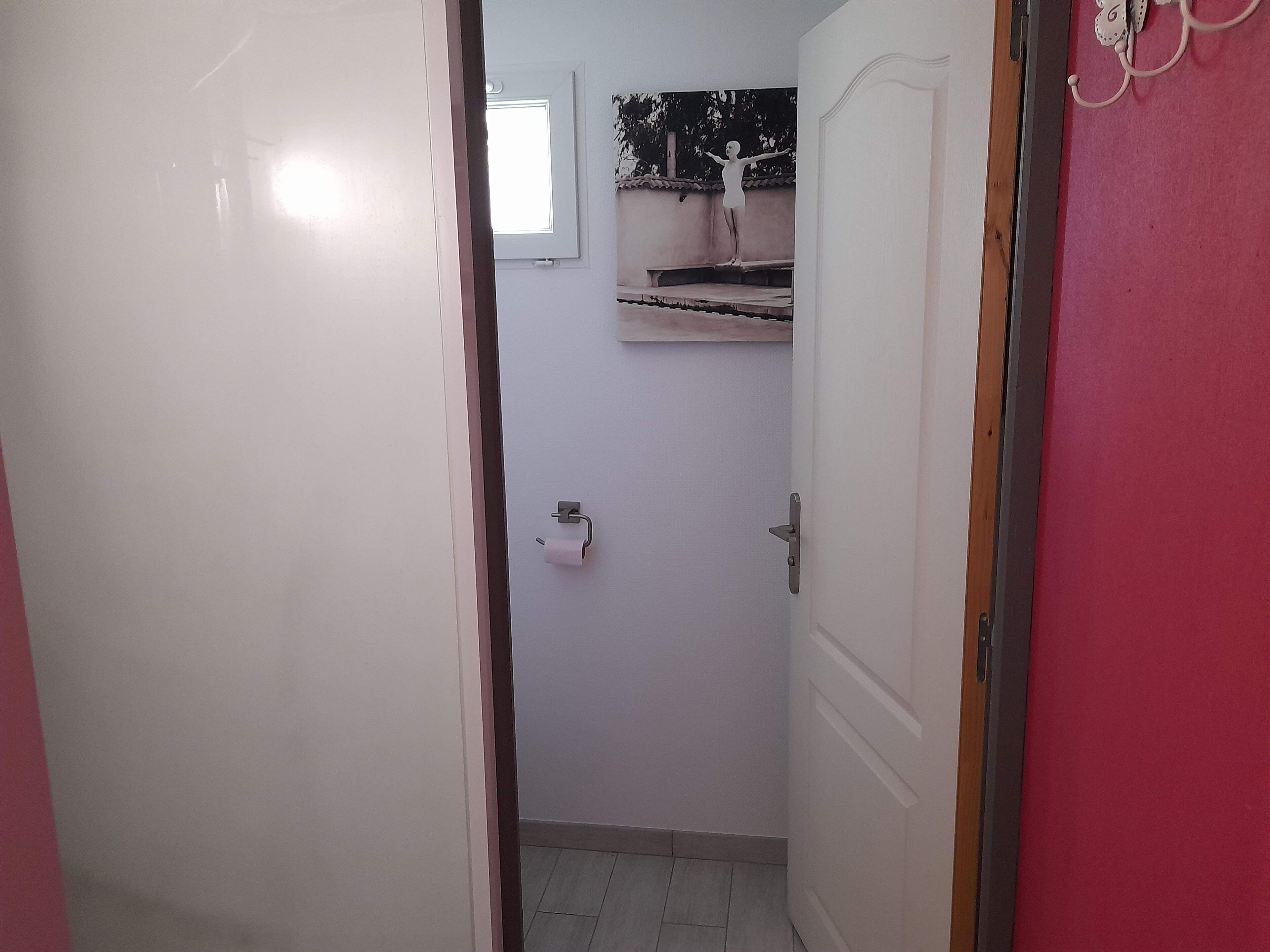 Un couloir étroit mène à une salle de bain équipée de toilettes, d’un porte-serviettes et d’une petite fenêtre.