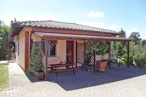 A small house with a tiled roof, a covered patio with a table and chairs, and a fenced garden area.
