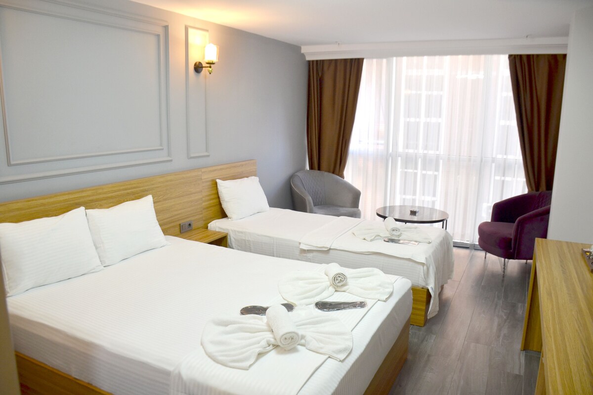 Ein Hotelzimmer mit zwei Betten, einem Sessel, einem kleinen Tisch und Blick ins Freie durch große Fenster.