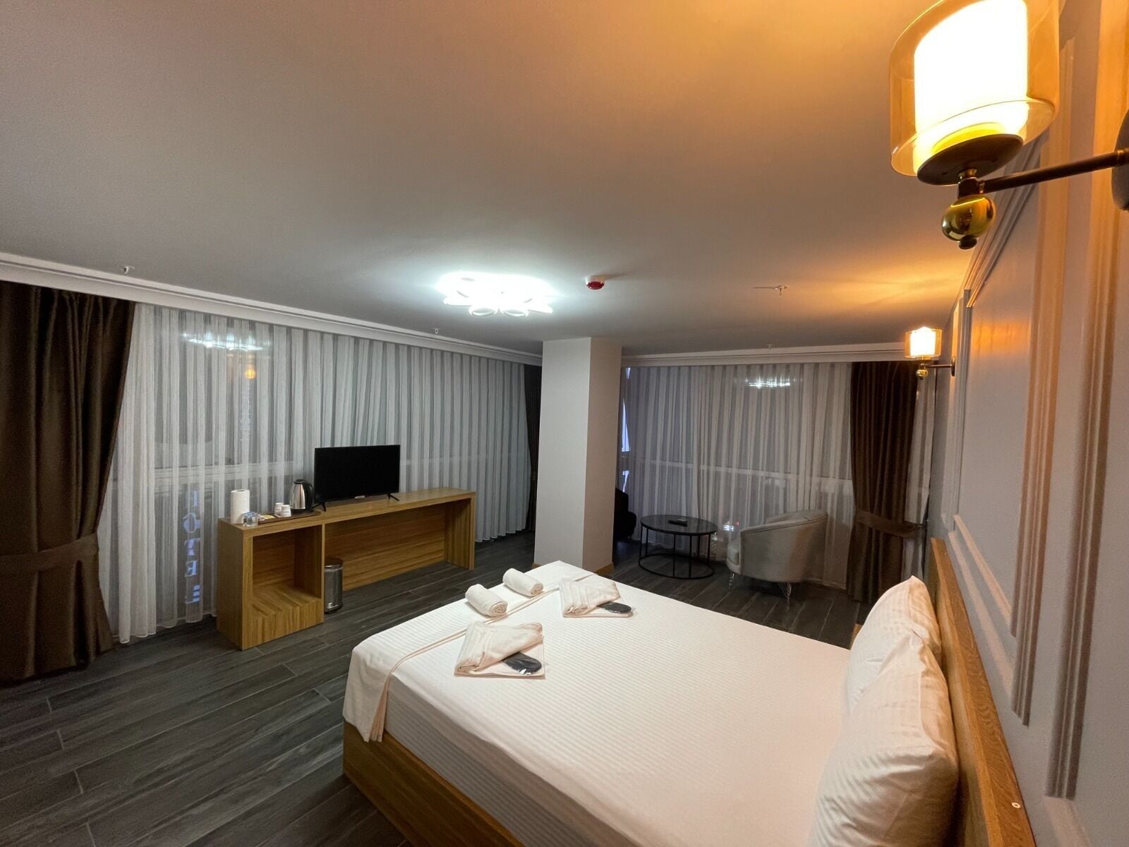 A hotel room with a bed, a TV on a wooden console, a desk, a chair, and a small table.