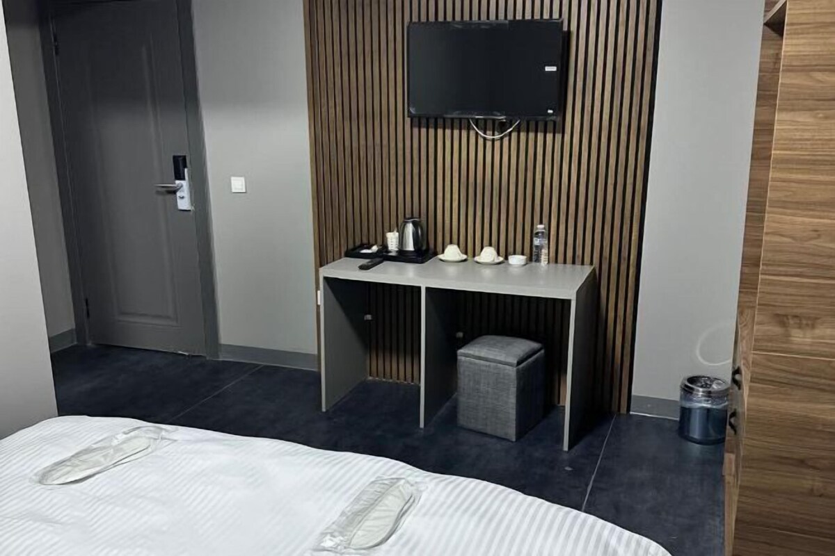 Ein Hotelzimmer mit Bett, Schreibtisch mit Fernseher, einem kleinen Hocker, einem Mülleimer und einer Tür.