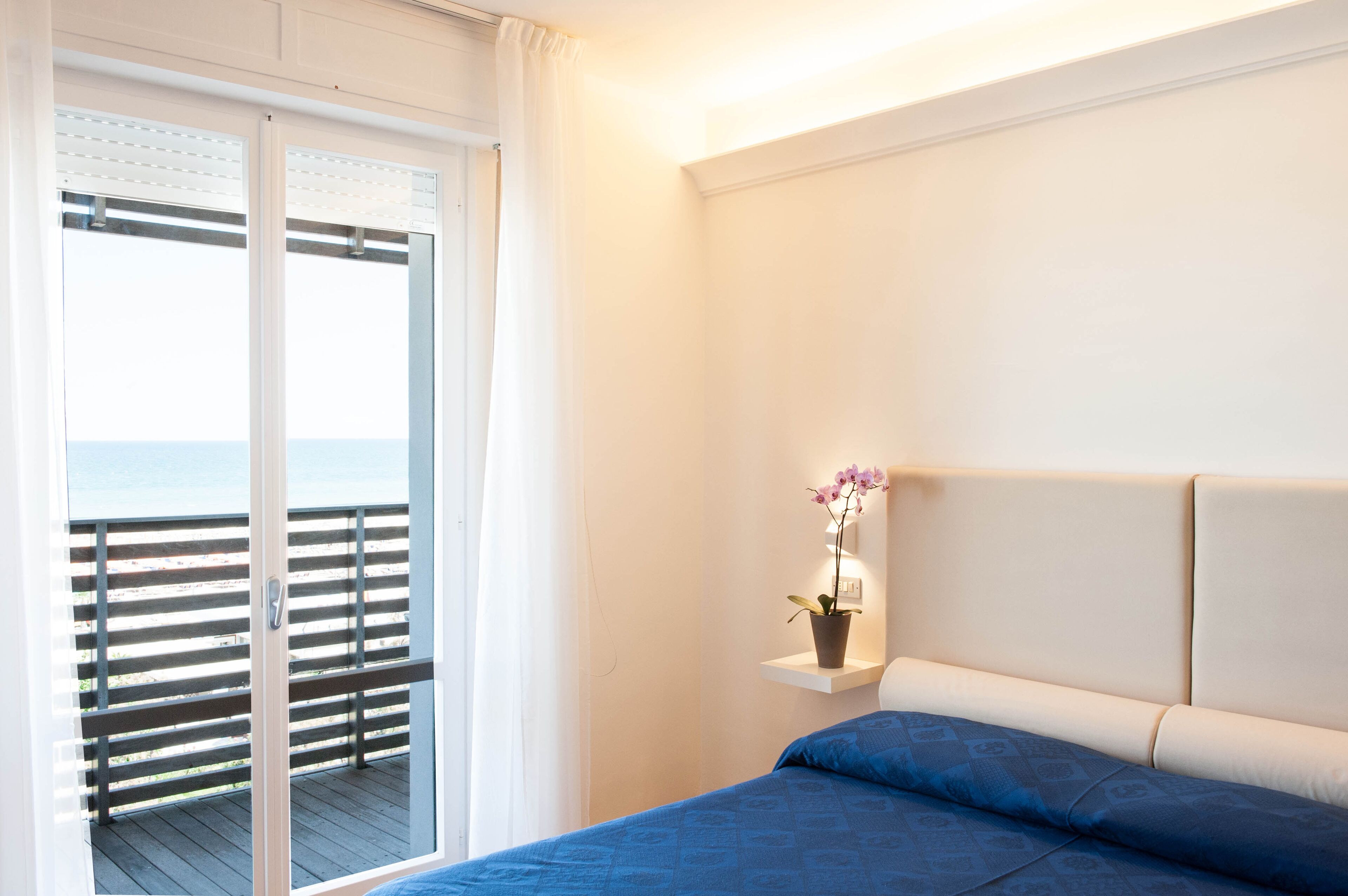 A bedroom with a bed, a nightstand with a potted plant, a window with a view of the sea, and a balcony with a railing.