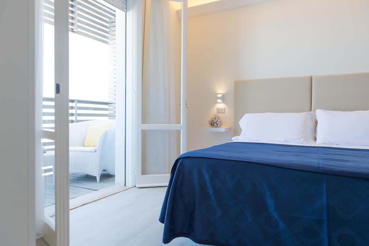Un dormitorio con una cama, una silla blanca, una mesita de noche y una lámpara de pared.