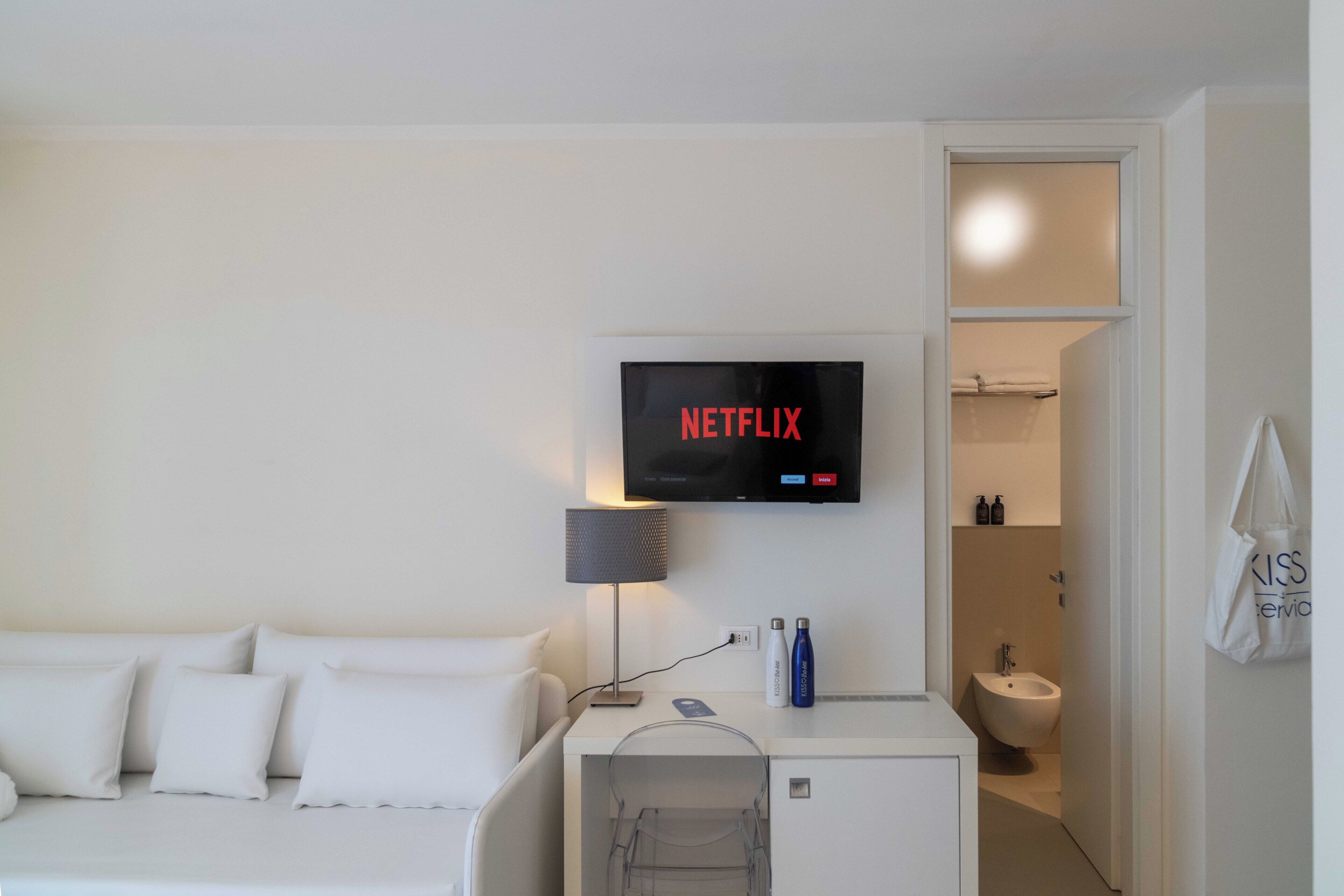 Una habitación de hotel con una cama, un escritorio con lámpara, un televisor que muestra 'NETFLIX', un baño con bidé y una toalla colgada en la pared.