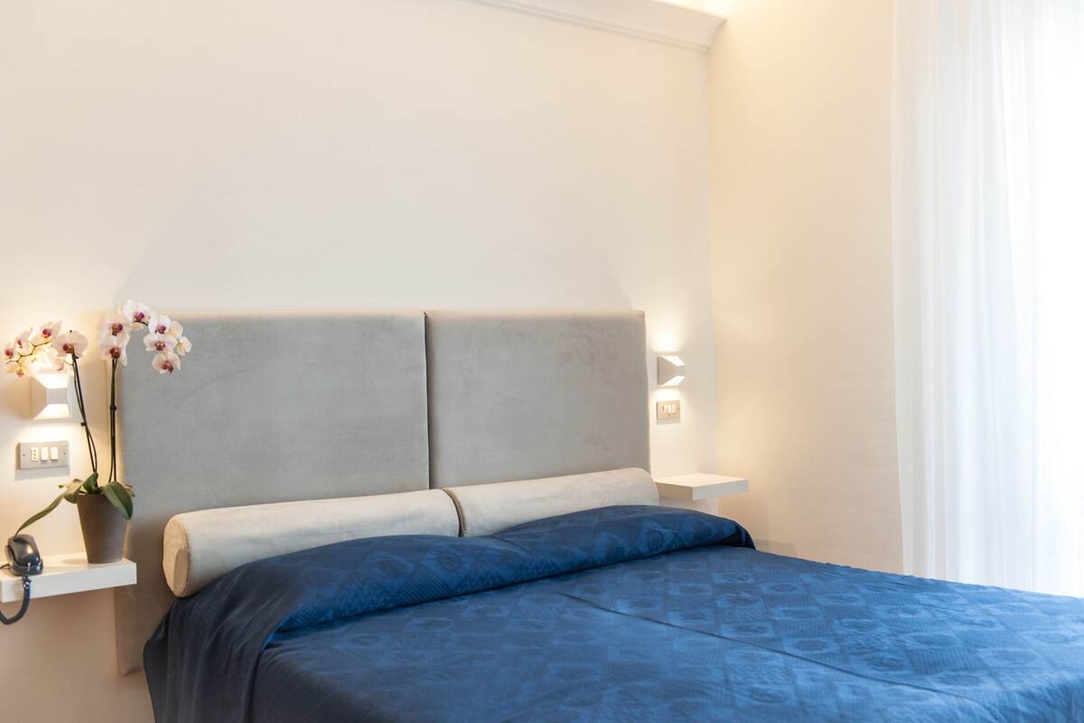 Una cama bien hecha con colcha azul y almohadas, cabecera gris y dos lámparas de noche.