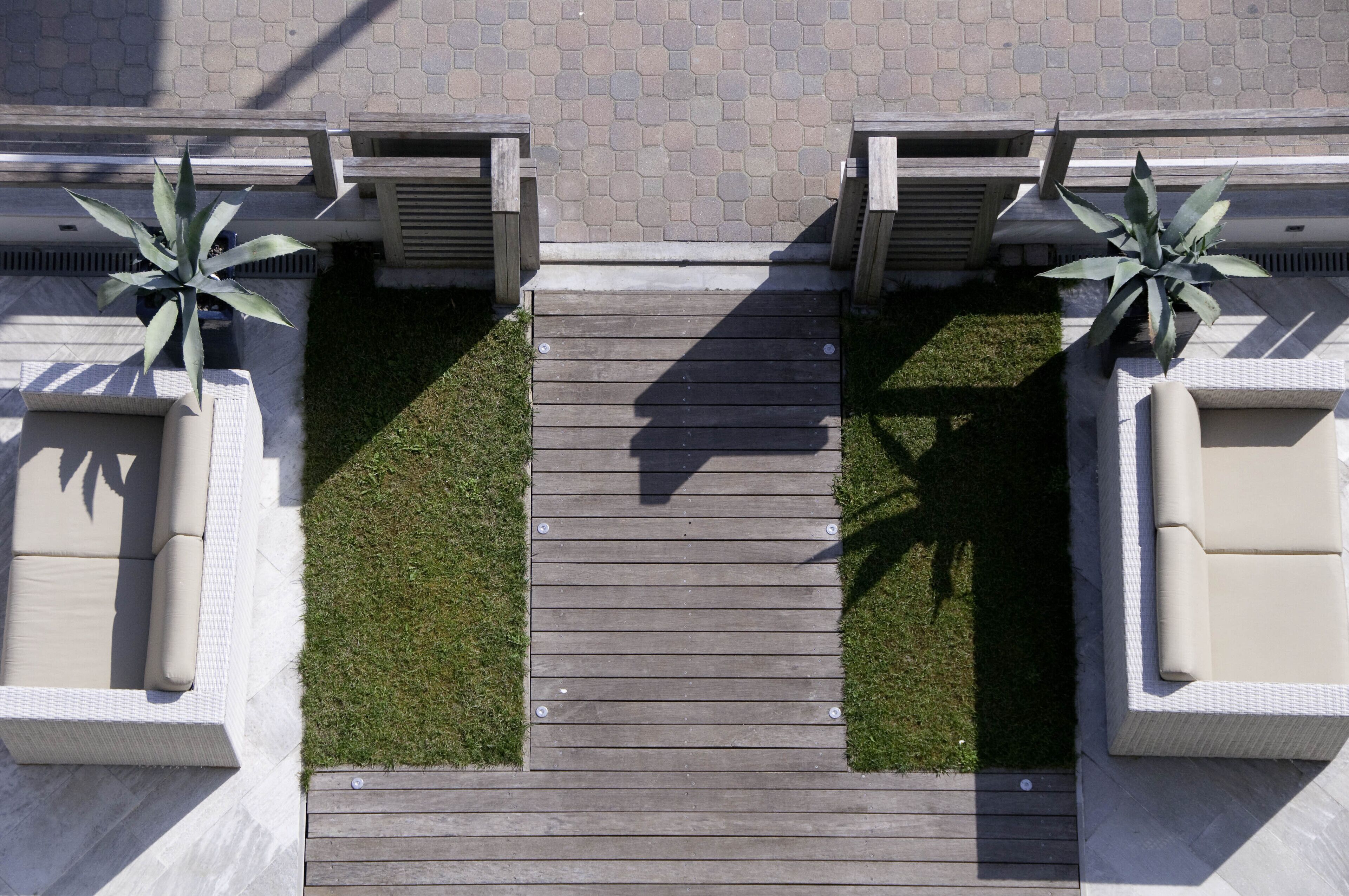 A wooden deck with a central pathway flanked by two planters with green plants, leading to a seating area with a sofa and a table.