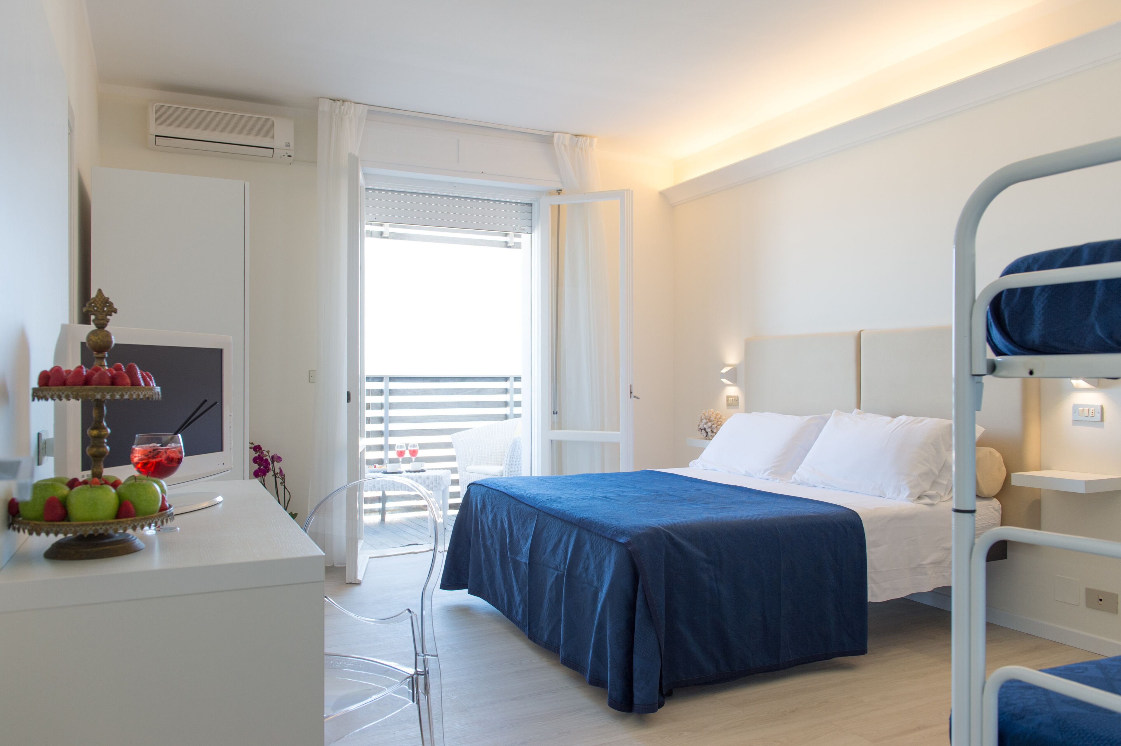 A hotel room with a bed, a desk with a fruit bowl, a chair, and a balcony with a view.