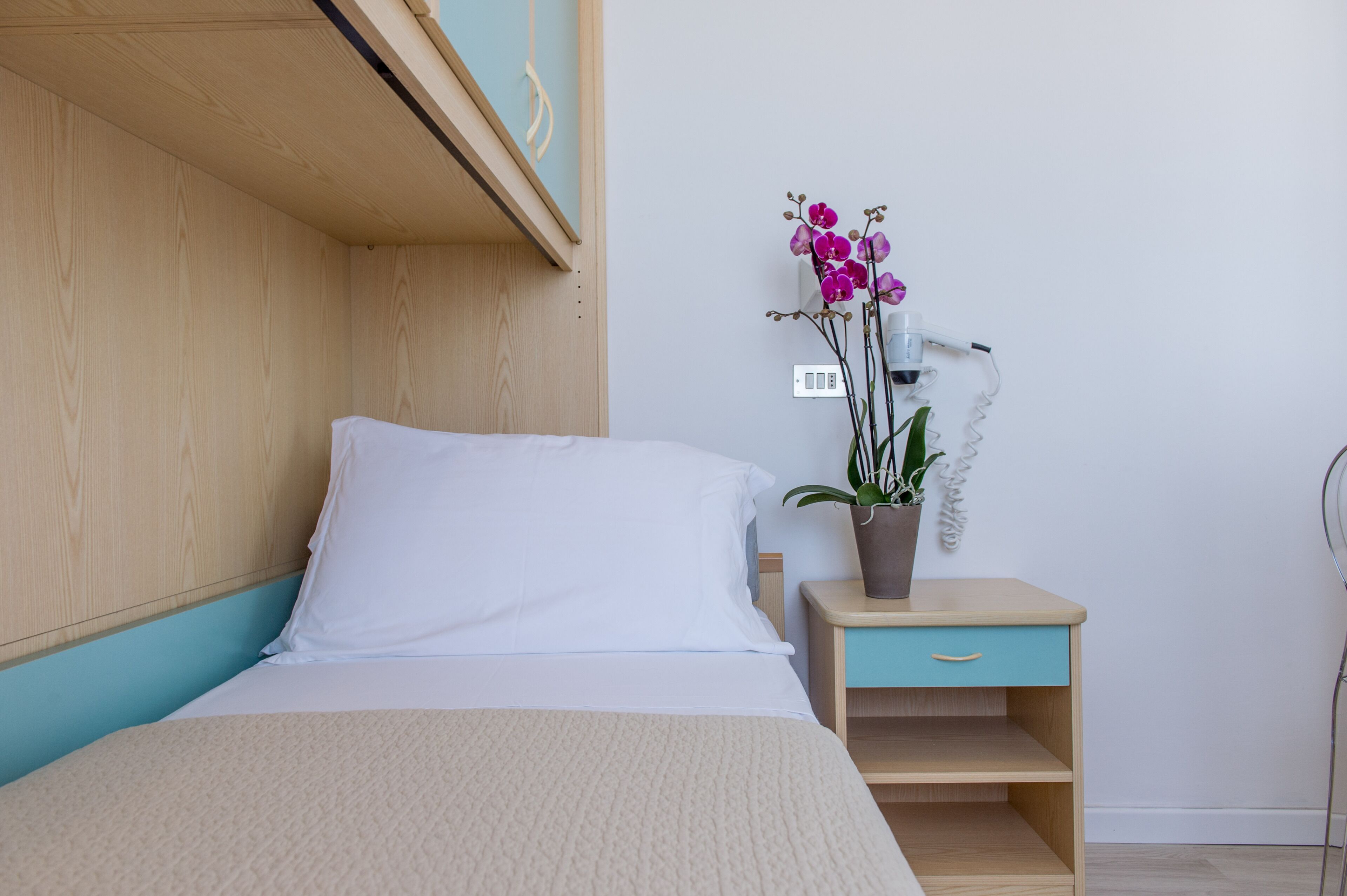 A neatly made bed with white bedding, a wooden headboard, a small bedside table with a drawer, and a potted orchid plant.