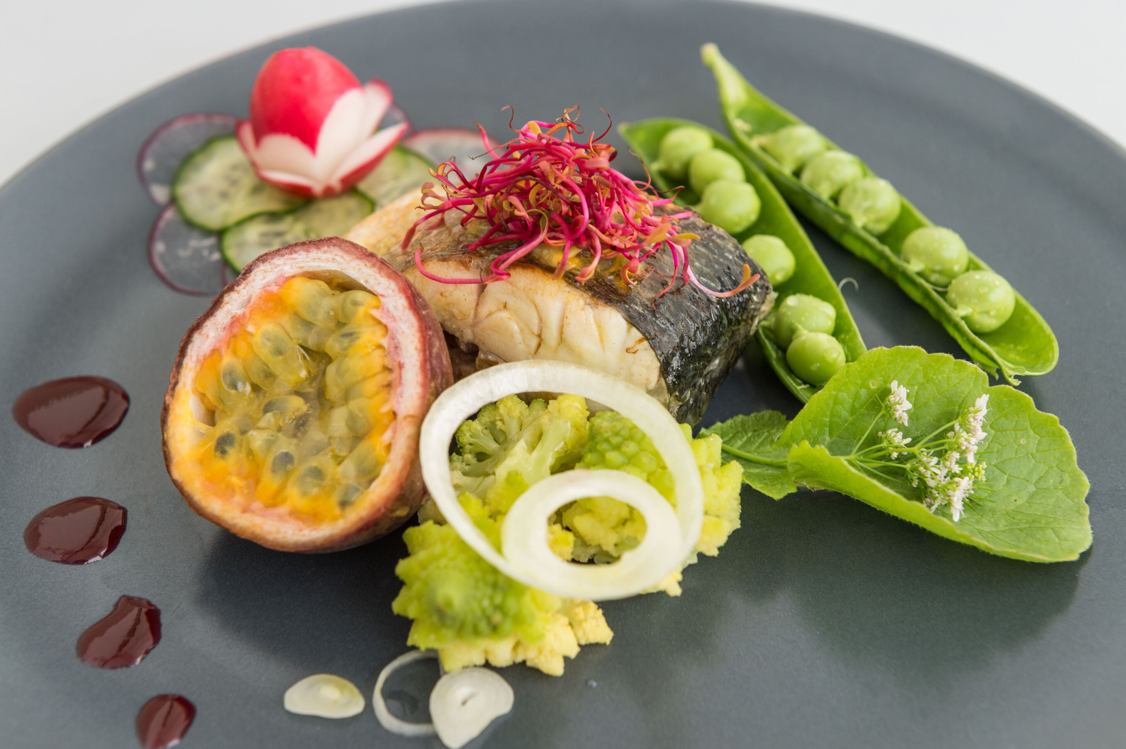 Une assiette contenant une variété de légumes et un morceau de poisson, ainsi que des tranches de fruit de la passion et des petits pois.