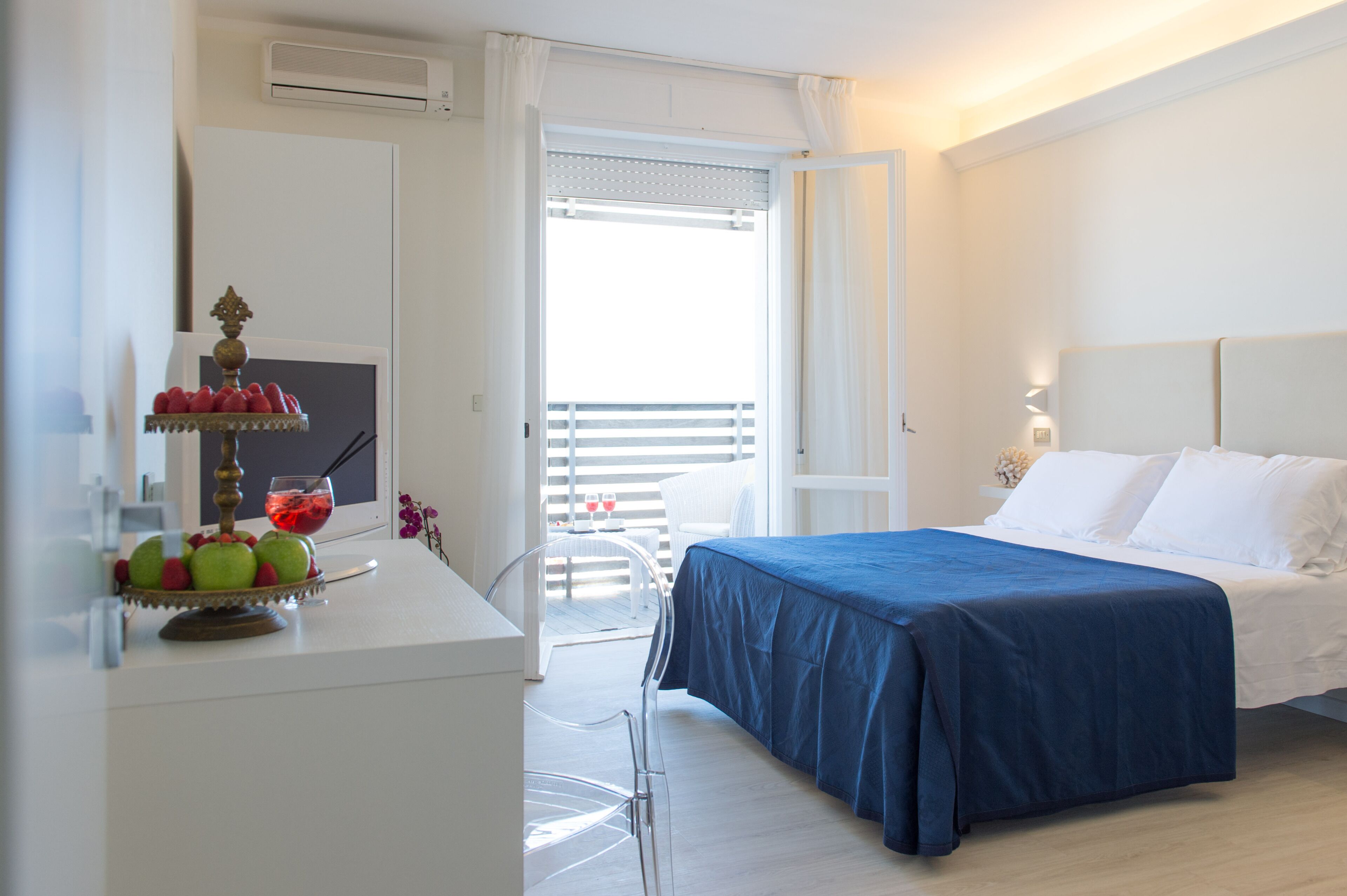 A hotel room with a bed, a desk with fruit and a drink, a balcony with a chair, and a white wall with a light fixture.