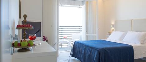 A hotel room with a bed, a desk with fruit and a drink, a balcony with a chair, and a white wall with a light fixture.