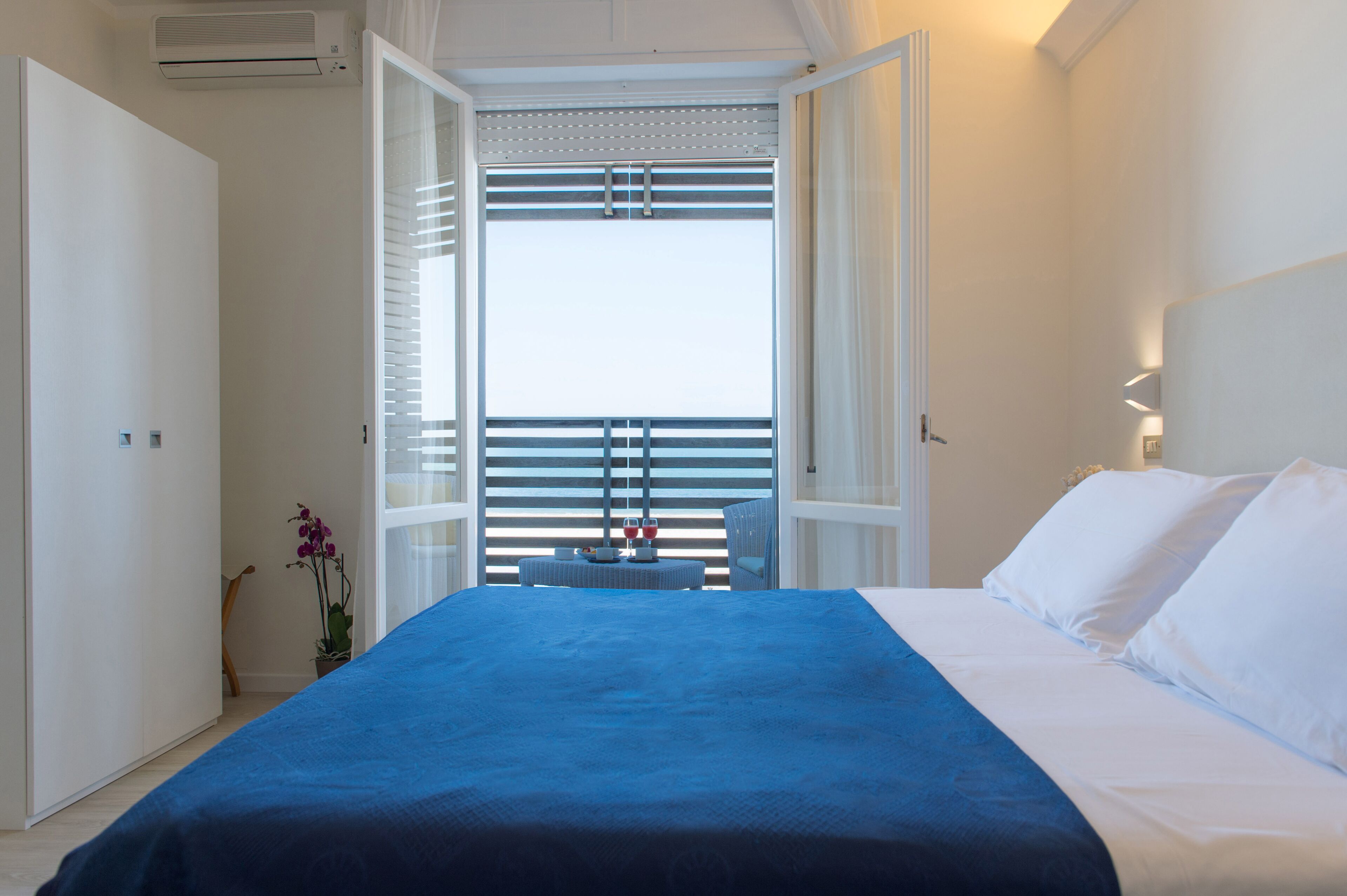 A bedroom with a bed, a nightstand, a potted plant, and a view through a window with blinds.