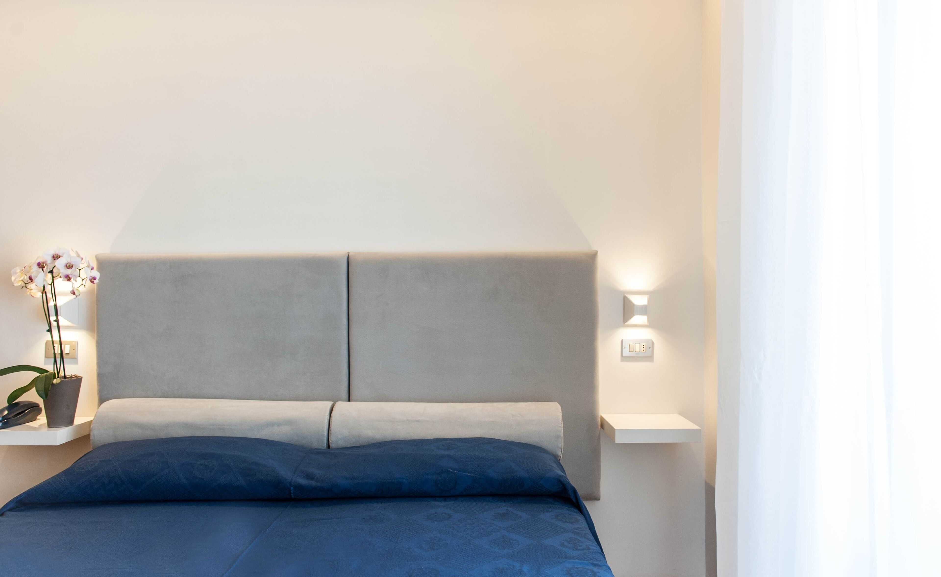 Una cama con cabecera gris, colcha azul y mesita de noche con una orquídea en maceta.