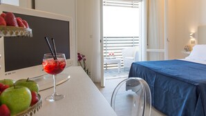 A hotel room with a bed, a desk with a computer monitor, a glass of red drink, and a bowl of fruit. - Kiss Beachfront Hotel & Restaurant (Cervia)