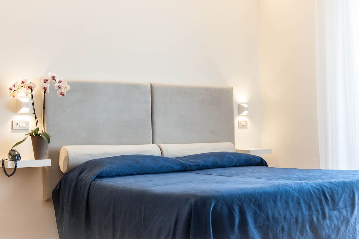 Una cama bien hecha con una colcha azul, un cabecero gris y dos lámparas de noche.