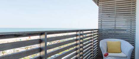 A balcony with a view of the beach, a wicker chair with a yellow cushion, and a small table with a drink and snacks.