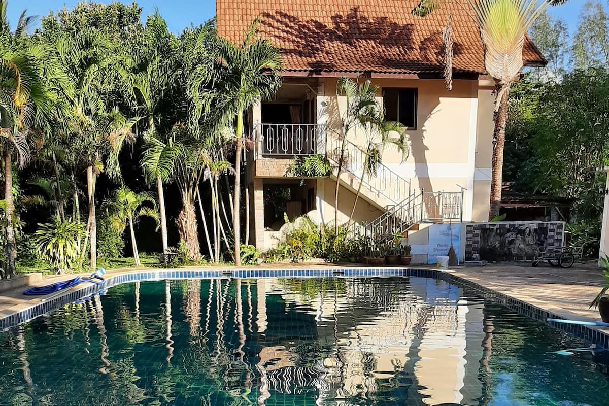 A swimming pool with clear blue water, surrounded by tropical plants and a building with a tiled roof.