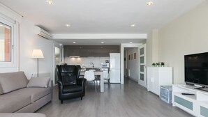 A modern living room with a sofa, a leather chair, a dining area, and a kitchen in the background. - UHC SOROLLA APARTMENTS (Salou)