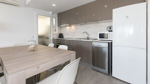 A modern kitchen with a wooden dining table, white chairs, stainless steel appliances, and a white refrigerator. - UHC SOROLLA APARTMENTS (Salou)