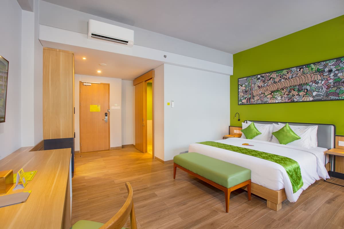 Quarto de hotel com piso de madeira, cama com roupa de cama verde e branca, escrivaninha com cadeira e uma pintura grande na parede.