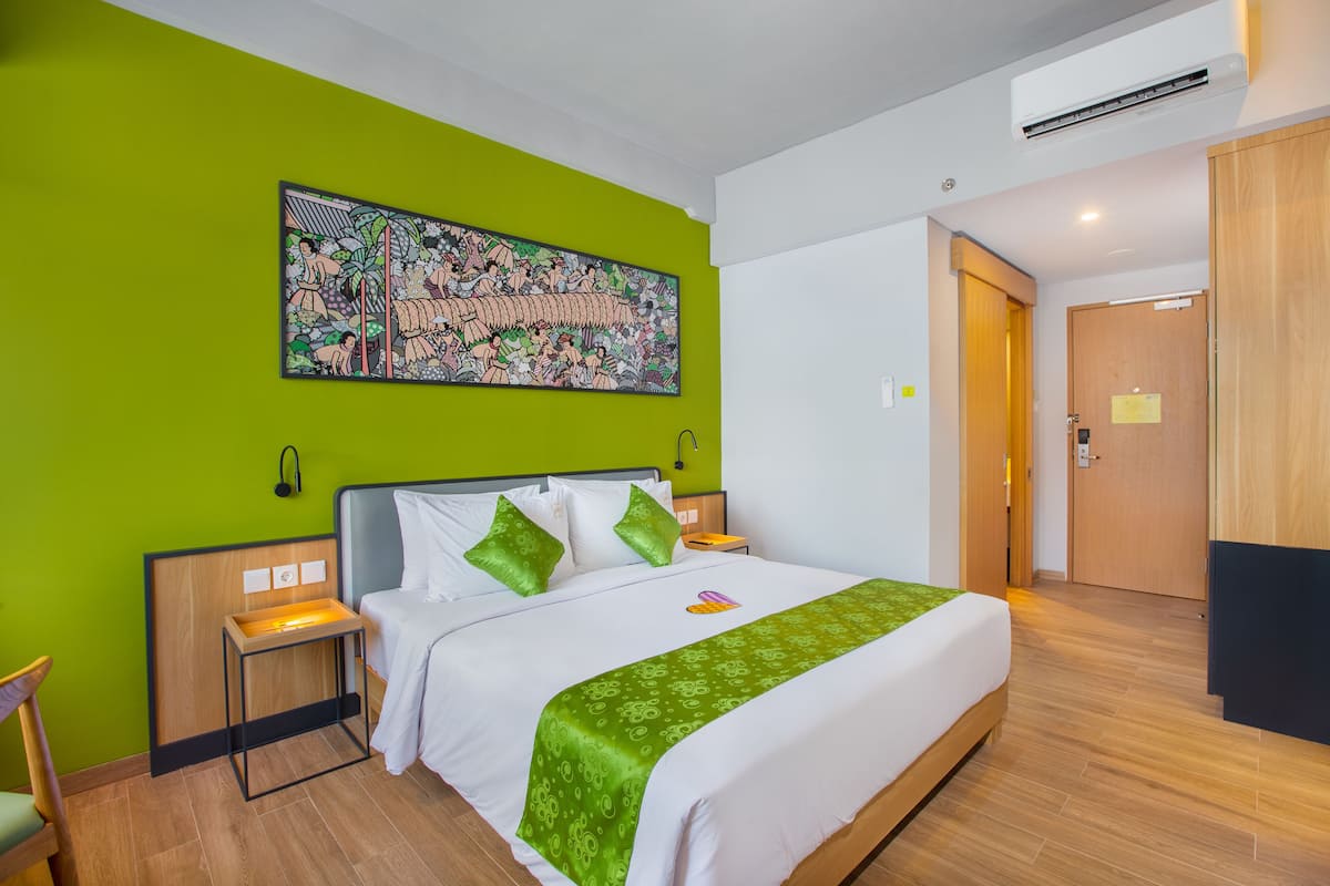 Quarto de hotel com uma cama grande, paredes verdes, uma obra de arte colorida sobre a cama, uma porta de madeira e uma pequena mesa de cabeceira com abajur.
