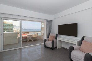 A modern hotel room with a balcony, a flat-screen TV, and a seating area with two armchairs.